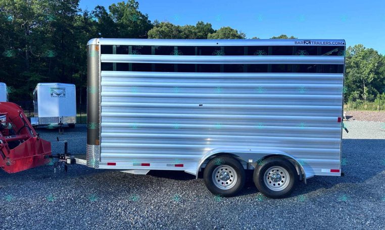 Silver aluminum livestock trailer parked on a gravel lot with a row of trees in the background.