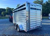 Silver aluminum livestock trailer on a gravel lot with a red tractor nearby and trees in the background, under a blue sky with sunlight.