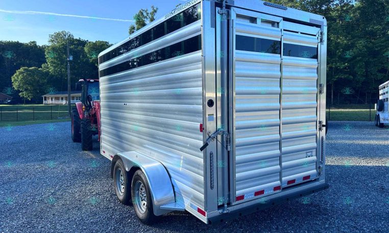 Silver aluminum livestock trailer on a gravel lot with a red tractor nearby and trees in the background, under a blue sky with sunlight.