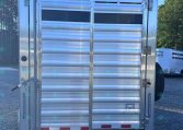 Silver aluminum trailer rear with vertical latch, roll-up gate, and safety latches in a gravel lot; other trailers visible in background.