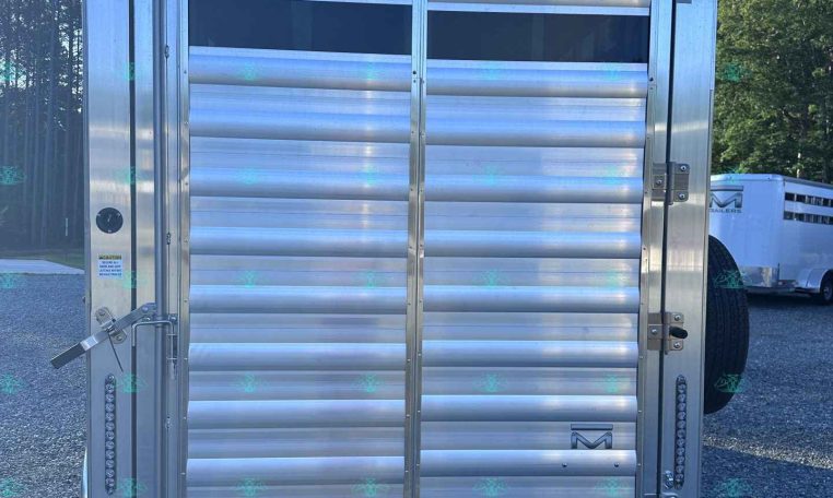 Silver aluminum trailer rear with vertical latch, roll-up gate, and safety latches in a gravel lot; other trailers visible in background.