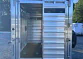 Rear view of an open aluminum livestock trailer with the doors swung wide, showing an empty interior on a gravel lot.