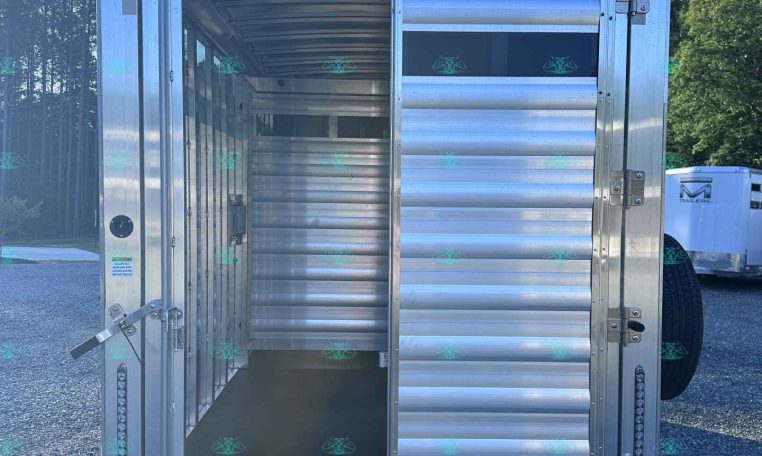 Rear view of an open aluminum livestock trailer with the doors swung wide, showing an empty interior on a gravel lot.