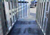Interior of a shiny aluminum horse trailer with divided stalls and open rear door, sunlight outside.