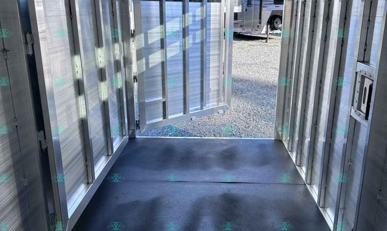 Interior of a shiny aluminum horse trailer with divided stalls and open rear door, sunlight outside.
