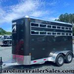 Black horse trailer with multiple horizontal windows, parked in a gravel lot with other trailers and trees in the background.