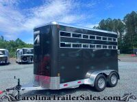 Black horse trailer with multiple horizontal windows, parked in a gravel lot with other trailers and trees in the background.