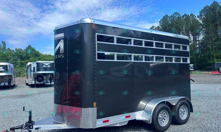 Black horse trailer with multiple horizontal windows, parked in a gravel lot with other trailers and trees in the background.