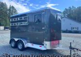 Dark blue horse trailer with windows and dual axles parked on a gravel lot, trees in the background and a building nearby.