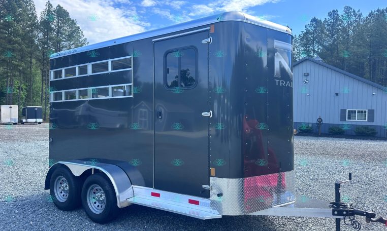 Dark blue horse trailer with windows and dual axles parked on a gravel lot, trees in the background and a building nearby.