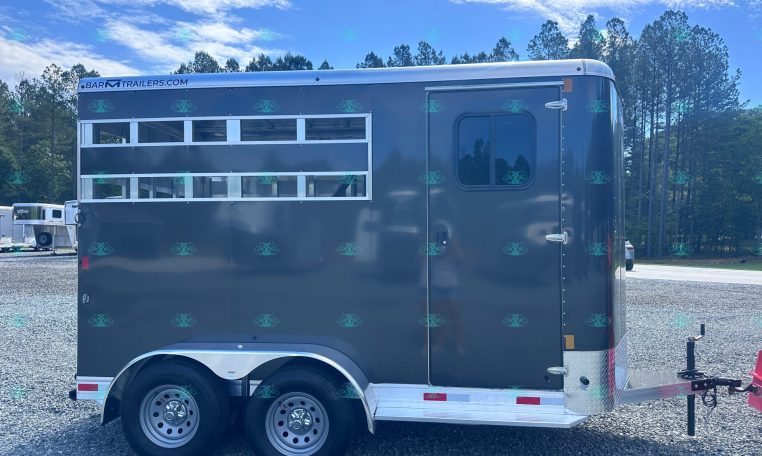Side view of a dark gray horse trailer with windows and a door, parked on a gravel lot with trees in the background; branding along the top edge reads Bar M Trailers.com.