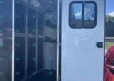 Open white horse trailer showing partitioned stalls and wall-mounted hooks inside, with door ajar against a blue sky backdrop.