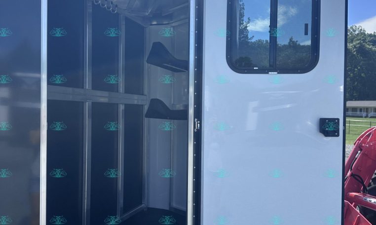 Open white horse trailer showing partitioned stalls and wall-mounted hooks inside, with door ajar against a blue sky backdrop.
