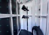 Interior of a metal trailer showing black storage panels, wall hooks, and a mounted spare tire on the floor near the back door area