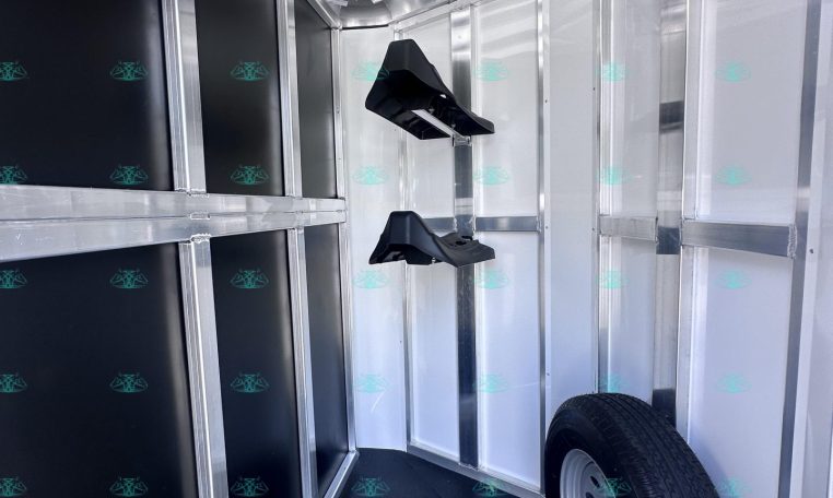 Interior of a metal trailer showing black storage panels, wall hooks, and a mounted spare tire on the floor near the back door area