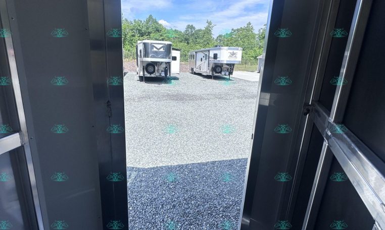 Interior of a metal trailer with the rear door open, revealing two horse trailers parked on a gravel lot under a blue sky.