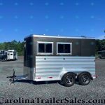 Black and silver trailer with two windows on a gravel lot, surrounded by other trailers under a clear blue sky.