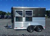 Silver and dark gray trailer with two silver-framed barred windows, hitch at the front, parked on a gravel lot with other trailers under a blue sky.