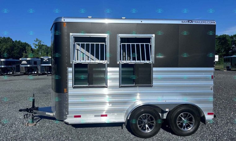 Silver and dark gray trailer with two silver-framed barred windows, hitch at the front, parked on a gravel lot with other trailers under a blue sky.