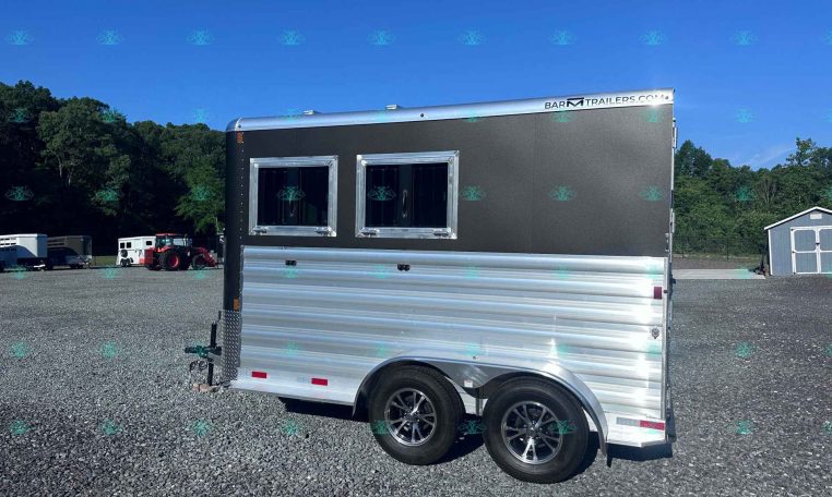 Black and silver two-window trailer parked on a gravel lot with a blue sky and trees in the background.