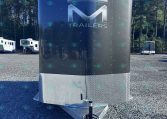 Front view of a large dark trailer with a metallic M logo and the word TRAILERS, parked on a gravel lot with trees in the background.