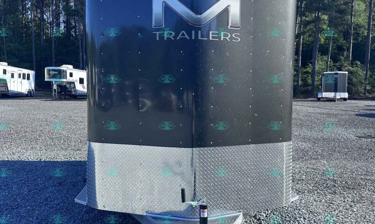 Front view of a large dark trailer with a metallic M logo and the word TRAILERS, parked on a gravel lot with trees in the background.