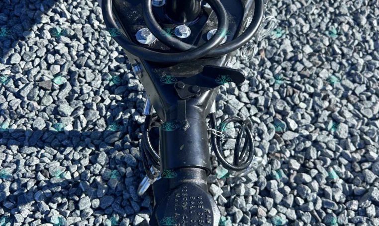Close-up of a trailer hitch with a vertical jack, safety chains, and coiled cables on gray gravel.