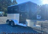 Black tandem-axle enclosed trailer with diamond-plate front right next to a hitch, parked on a gravel lot with trees in the background