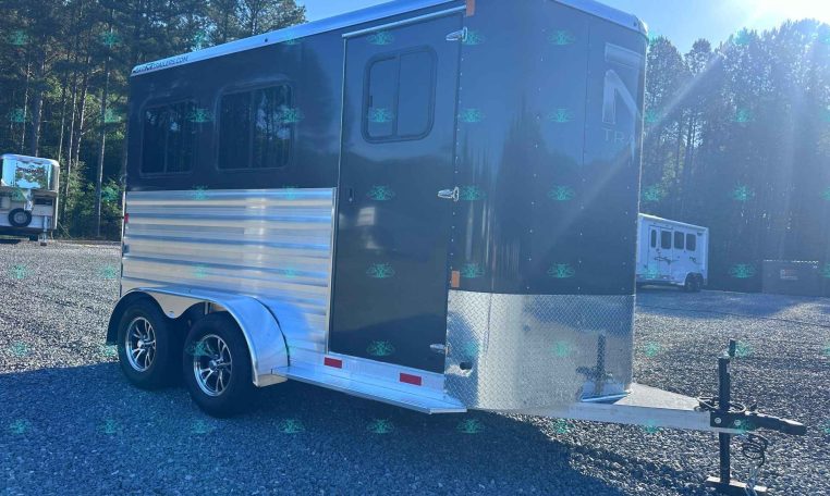 Black tandem-axle enclosed trailer with diamond-plate front right next to a hitch, parked on a gravel lot with trees in the background