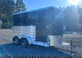 Blue horse trailer in a gravel lot with evergreen trees and bright sun glare in the background.