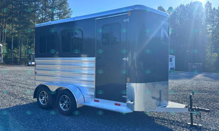 Blue horse trailer in a gravel lot with evergreen trees and bright sun glare in the background.