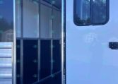 Rear-opening of a white horse trailer, showing an empty aluminum stall compartment with a row of hooks along the ceiling and a window on the side door