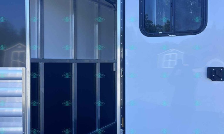 Rear-opening of a white horse trailer, showing an empty aluminum stall compartment with a row of hooks along the ceiling and a window on the side door
