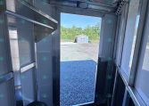 Interior of a metal trailer showing exposed framing and a spare tire, with the rear door open to a gravel lot and a small shed in the distance outside.