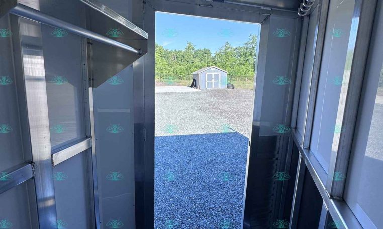 Interior of a metal trailer showing exposed framing and a spare tire, with the rear door open to a gravel lot and a small shed in the distance outside.