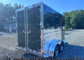 Rear view of a large black aluminum trailer with double doors, parked on a gravel lot beside a small building and trees under a blue sky.