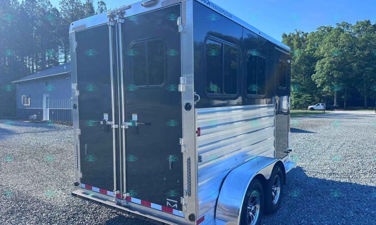 Rear view of a large black aluminum trailer with double doors, parked on a gravel lot beside a small building and trees under a blue sky.