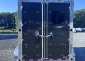 Rear view of a dark gray enclosed trailer with double doors and metal latches on a gravel lot, blue sky and trees in the background,