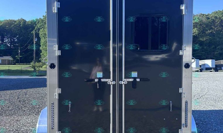 Rear view of a dark gray enclosed trailer with double doors and metal latches on a gravel lot, blue sky and trees in the background,