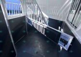 Interior of a aluminum horse trailer with divider gates and a small bench seat along the wall, sunlit window on left side