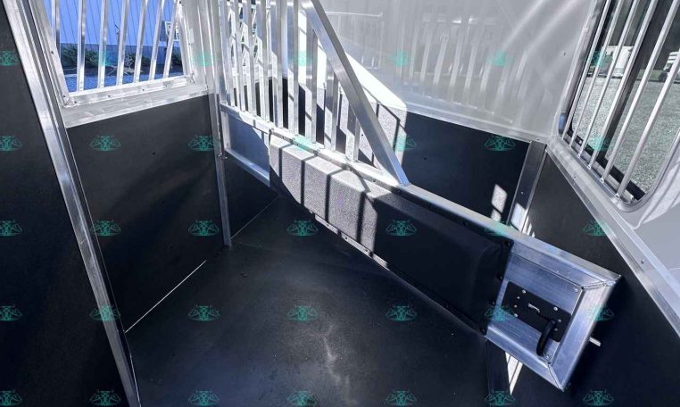 Interior of a aluminum horse trailer with divider gates and a small bench seat along the wall, sunlit window on left side