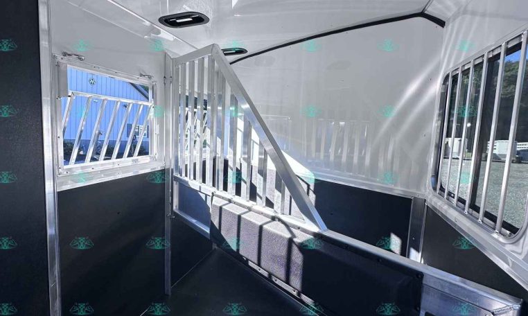 Interior of a white horse trailer with a partitioned stall, steps, and a side window showing outside equipment.