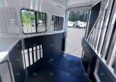 Interior of a metallic horse trailer with barred windows, dark rubber floor, and open exterior ramp showing other trailers outside