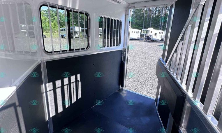 Interior of a metallic horse trailer with barred windows, dark rubber floor, and open exterior ramp showing other trailers outside