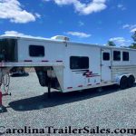 White two-axle gooseneck horse trailer parked on a gravel lot under a clear blue sky with trees in the background; red towing equipment in the foreground and a second trailer visible in the distance.