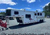 White two-axle gooseneck horse trailer parked on a gravel lot under a clear blue sky with trees in the background; red towing equipment in the foreground and a second trailer visible in the distance.