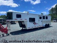 White two-axle gooseneck horse trailer parked on a gravel lot under a clear blue sky with trees in the background; red towing equipment in the foreground and a second trailer visible in the distance.