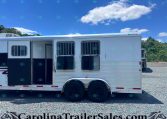 White horse trailer with open side doors and barred windows parked on a gravel lot under a bright blue sky with clouds.