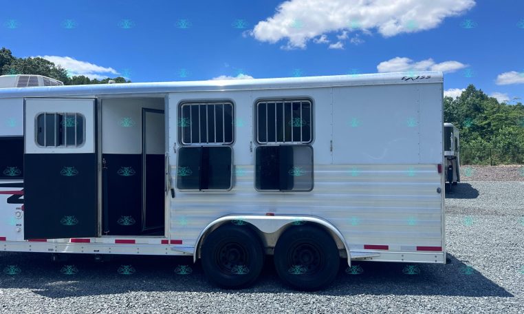 White horse trailer with open side doors and barred windows parked on a gravel lot under a bright blue sky with clouds.