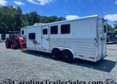White horse trailer with multiple windows parked on a gravel lot beside a red tractor, against trees and a blue sky.